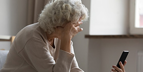 Elderly woman sitting on a sofa holding her head while holding a smart phone with the other hand